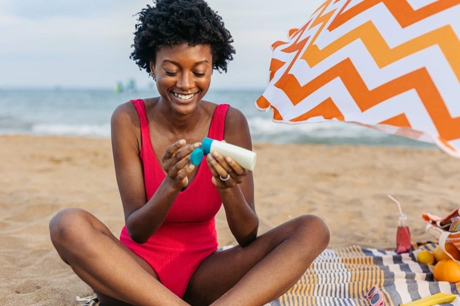 Women applying sunscreen while sitting on a beach beside a striped umbrella and picnic items