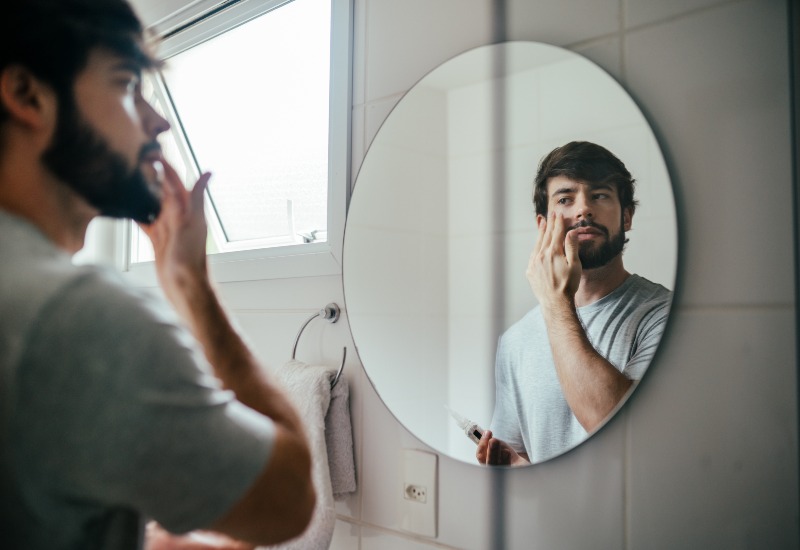 A man is applying skincare product in front of a bathroom mirror in Dallas, TX, and surrounding area